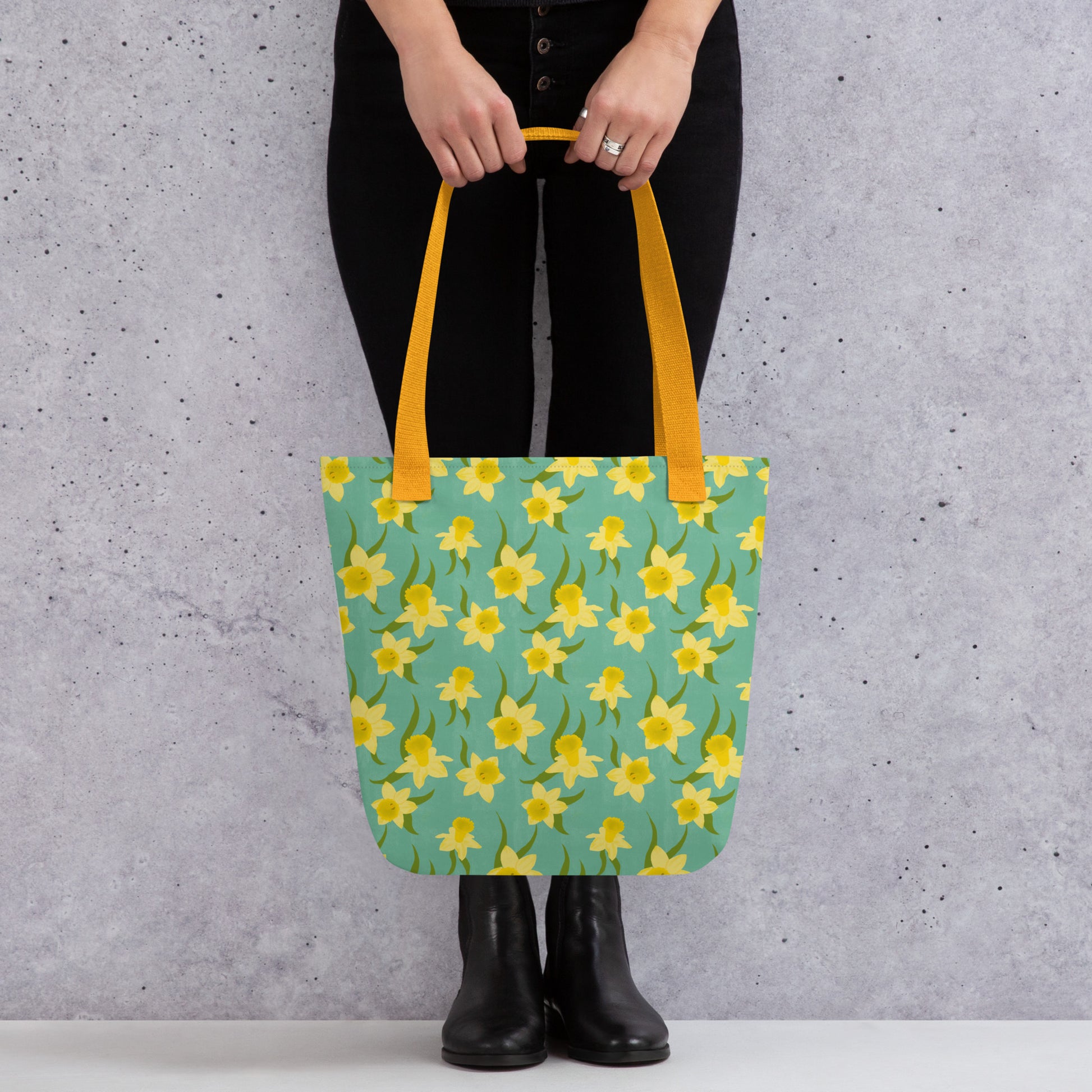Tote bag with yellow daffodil floral pattern by Carmel Rimmer and a yellow handle held by a person against a grey background