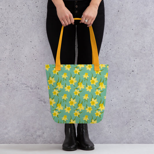 Tote bag with yellow daffodil floral pattern by Carmel Rimmer and a yellow handle held by a person against a grey background