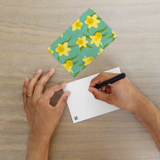 A person's hands are shown writing a message on a blank postcard with a floral daffodil patterned postcard by Carmel Rimmer lying flat next to it.