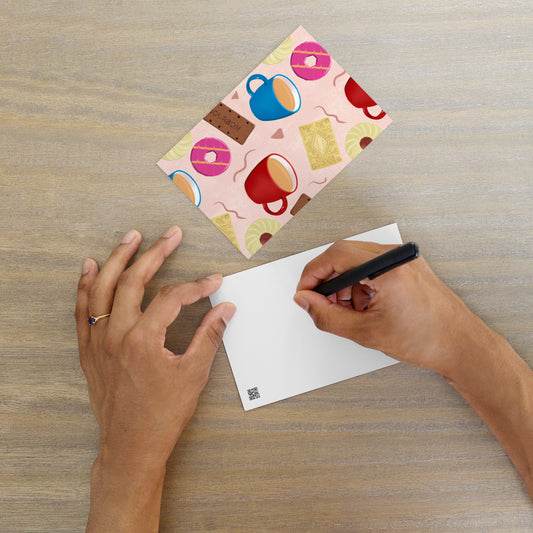 A person's hands are shown writing a greeting on a postcard with a 'Tea and Biscuits' pattern by Carmel Rimmer, placed on a wooden table.