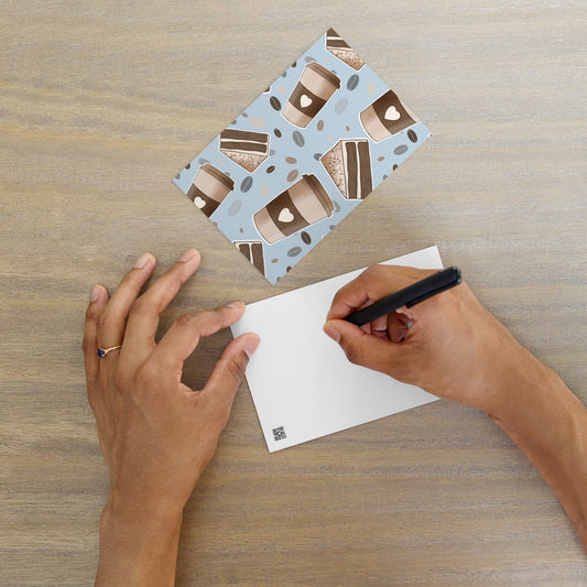 A person's hands are shown writing a message on a 'Coffee and Cake' patterned postcard designed by Carmel Rimmer. with a white matte back. The postcard has a blue background with illustrations of coffee cups and Chocolate cake slices.