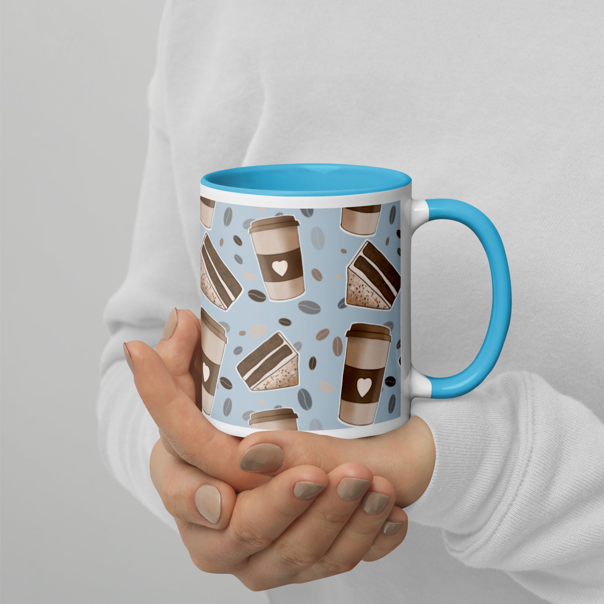 A ceramic mug with a 'Coffee & Cake' pattern by Carmel Rimmer and a blue rim and handle, being held in a person's hands.