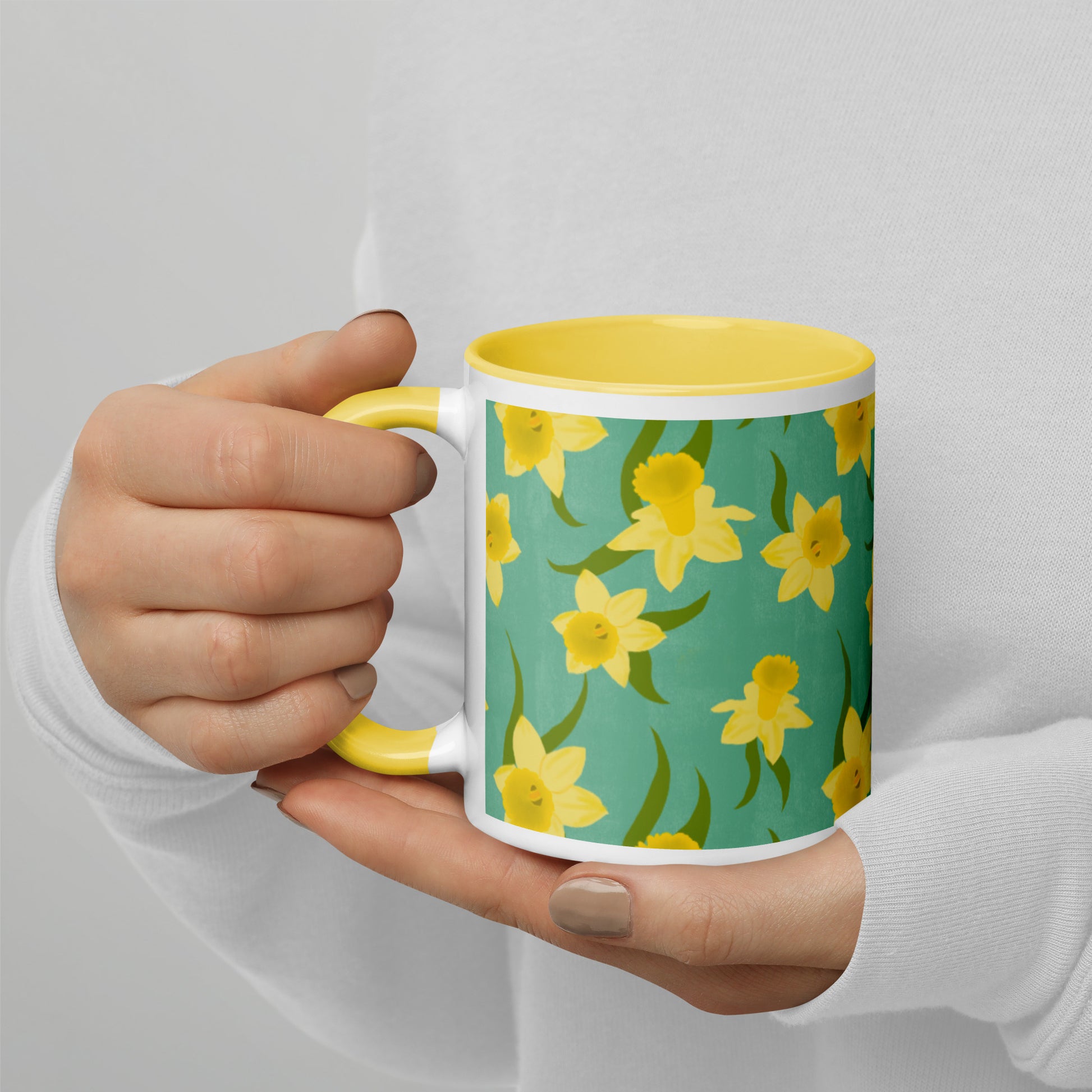 A person holding a ceramic mug with a pattern of yellow daffodils on a teal background, and a yellow rim and handle. Design by Carmel Rimmer