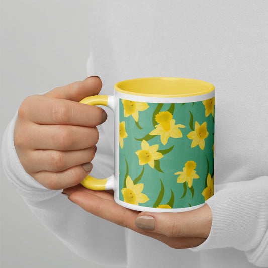 A person holding a ceramic mug with a pattern of yellow daffodils on a teal background, and a yellow rim and handle. Design by Carmel Rimmer
