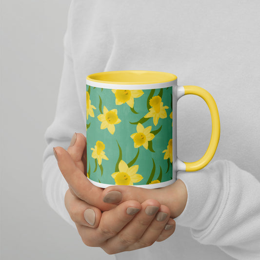 A person holding a ceramic mug with a pattern of yellow daffodils on a teal background, and a yellow rim and handle. Design by Carmel Rimmer